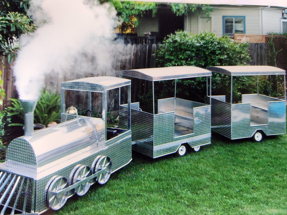 A small metal train with two open passenger cars on a lawn, smoke rising from the stack